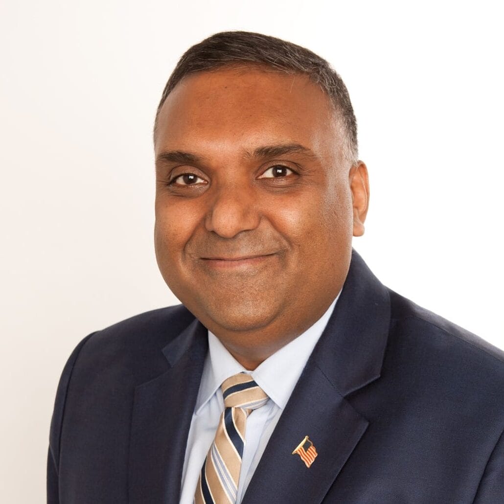 A man in a navy blue suit, light blue shirt, and striped tie smiles at the camera. He has short dark hair, wears a small U.S. flag pin on his lapel, and is a member of the MassArt Board of Trustees against a plain white background.