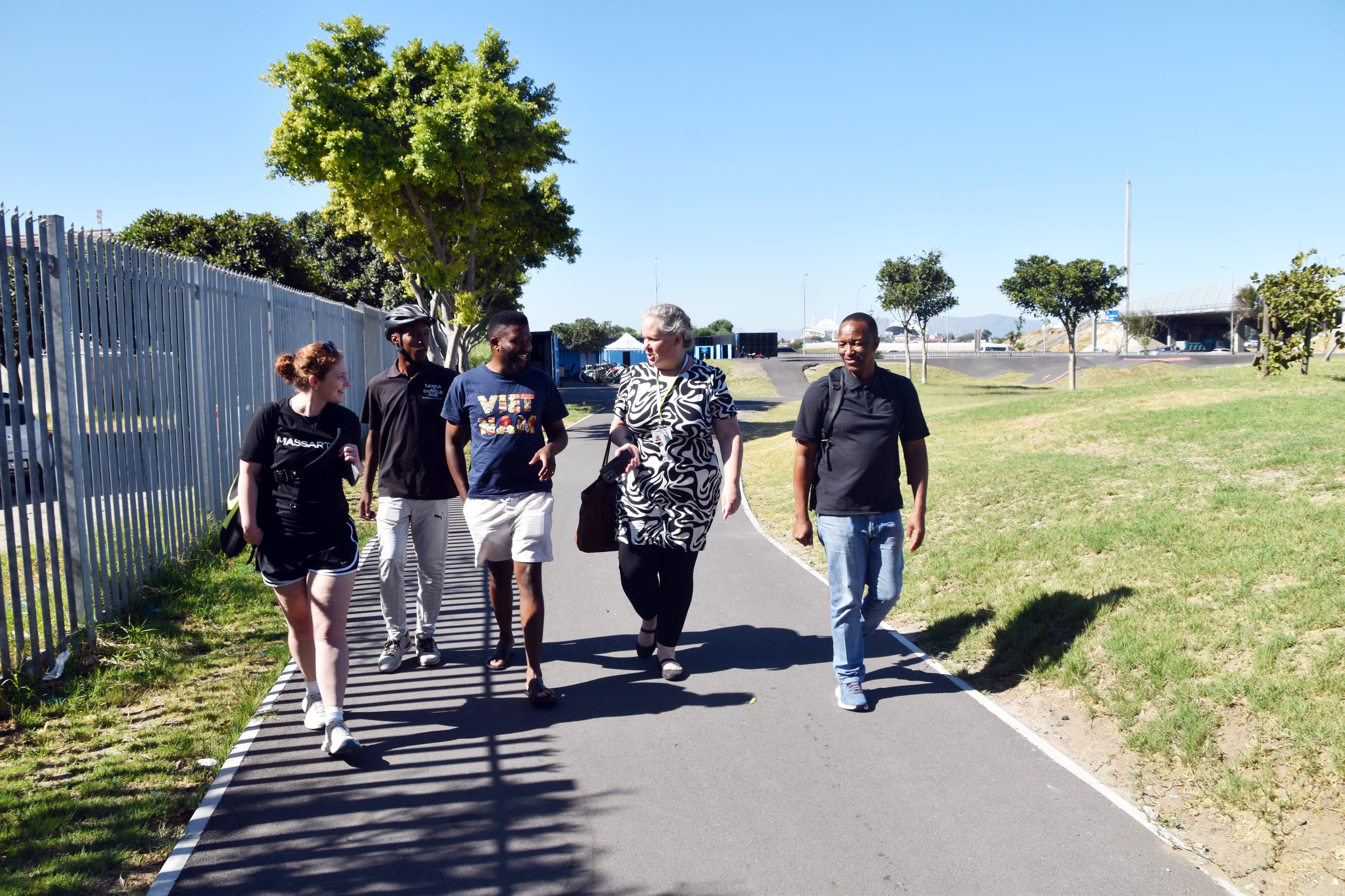 Five people walk together on a paved path beside Reliable Building and a grassy area in Langa, South Africa, under a clear blue sky. Engaged in conversation, they pass trees and distant structures visible in the background.