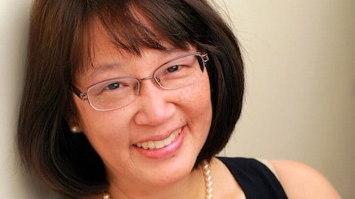 A woman with straight dark hair and glasses smiles at the camera. Wearing a sleeveless black top and a pearl necklace, this Common Good Award recipient stands confidently against a light-colored wall.