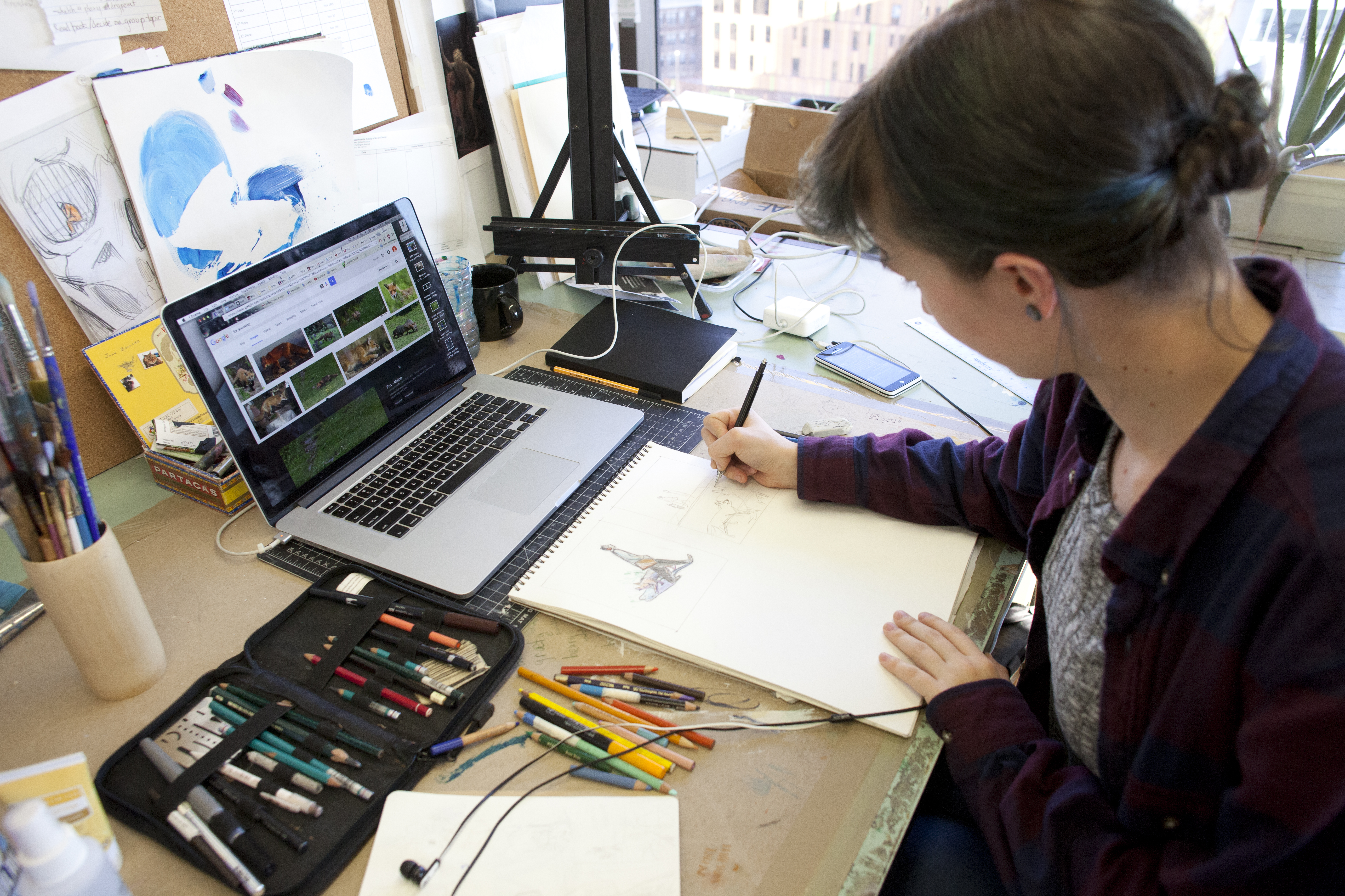 In a cozy studio, a person sits at a desk drawing in a sketchbook, surrounded by pens and art supplies, with their laptop open to animal reference images. Pre-semester energy fills the space, scattered papers and city views inspiring creativity.