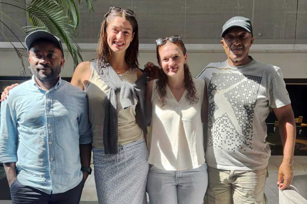 Four people stand side by side, smiling at the camera indoors in Langa, South Africa. Two women are in the middle, flanked by two men. Bright sunlight streams in, with a large leafy plant visible on the left.