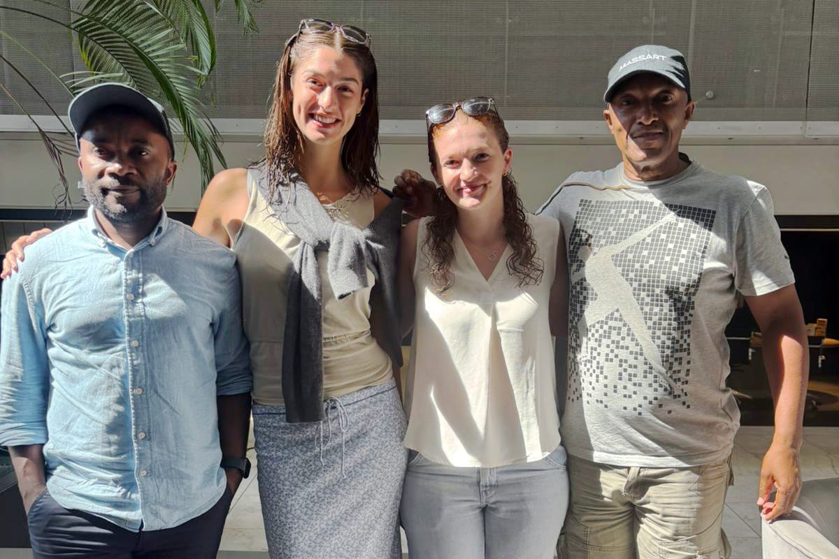 Four people stand side by side, smiling at the camera indoors in Langa, South Africa. Two women are in the middle, flanked by two men. Bright sunlight streams in, with a large leafy plant visible on the left.