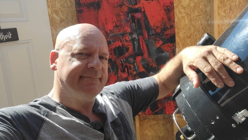 A man smiles at the camera while holding a large telescope, charting an out of this world moment. Behind him, a red and black abstract painting is mounted on a wooden wall as sunlight casts striking shadows across the scene.