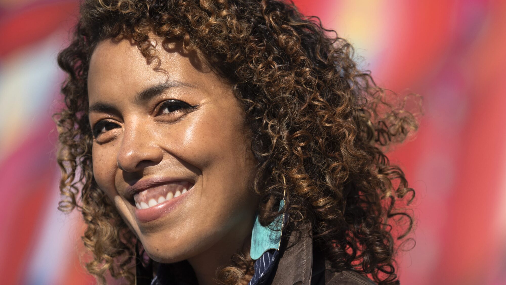 A woman with curly hair smiles while wearing a leather jacket and striped shirt, standing in front of a colorful, blurred background—one of the inspiring Common Good Award recipients.