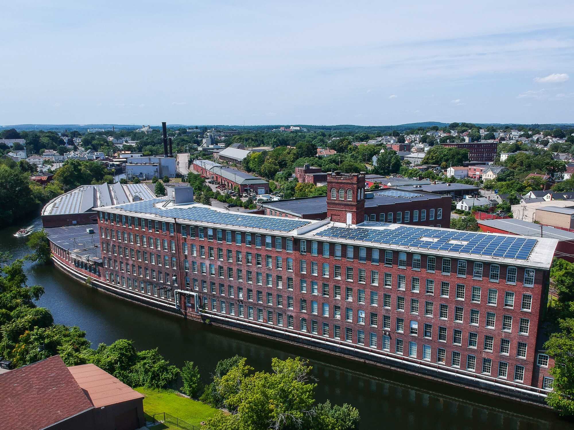 A large red brick mill building with many windows sits alongside a river, surrounded by trees and other industrial buildings under a blue sky. Solar panels cover much of its roof, reflecting its recognition among Common Good Award recipients.