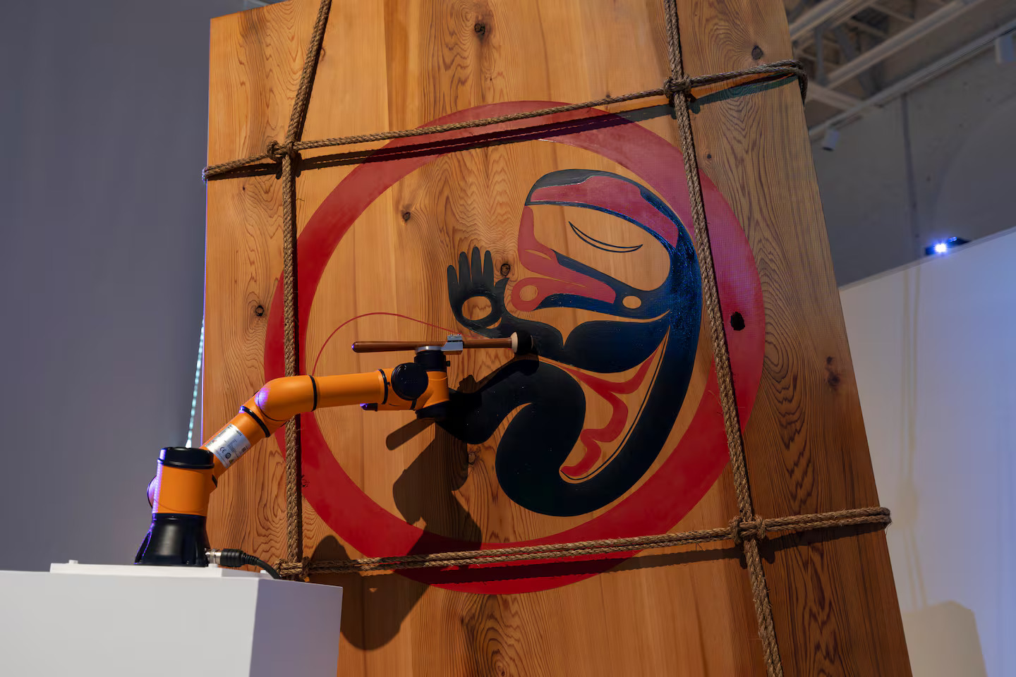 A robotic arm paints or engraves a stylized Indigenous art design of a human fetus on a large wooden panel, secured with rope. Created by an Indigenous artist, the piece is showcased in an exhibition space reminiscent of the Boston Triennial.