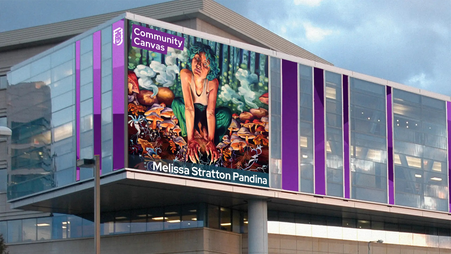 A digital display on a modern building shows mural artwork titled “Community Canvas” by alumna Melissa Stratton Pandina, featuring a colorful painting of a person surrounded by mushrooms. The building has purple and glass panels.