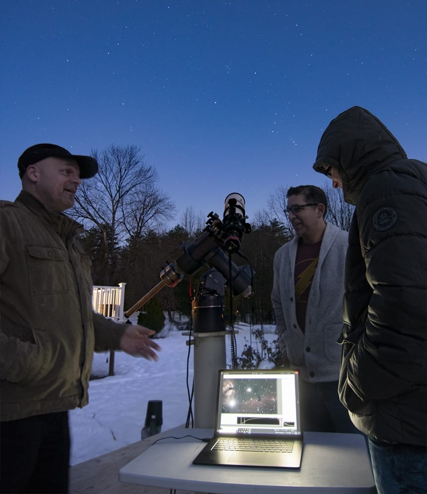 Three people stand outside at night in winter, gathered around a telescope and a laptop on a table. The laptop displays a star map, perhaps tracking an asteroid—a scene inspired by MassArt alum Sean Walker’s work with the International Astronomical Union.