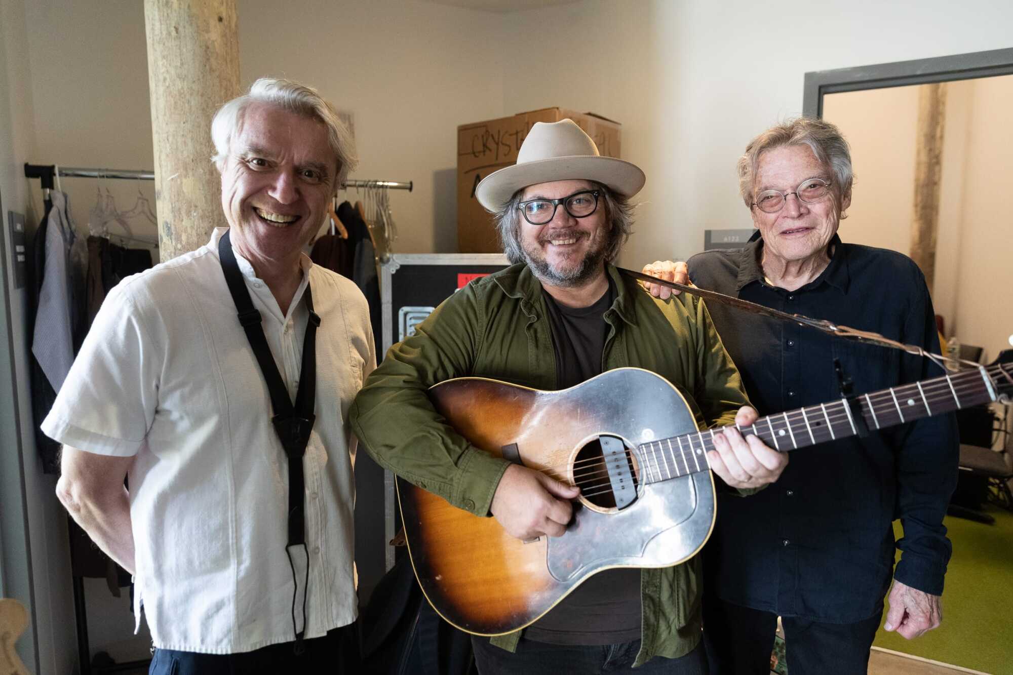 Three smiling men stand together indoors, appearing to be backstage. The man in the center, an Award Recipient, wears glasses and a hat while holding an acoustic guitar. The man on the left wears a white shirt and the rightmost man has on a dark vest.