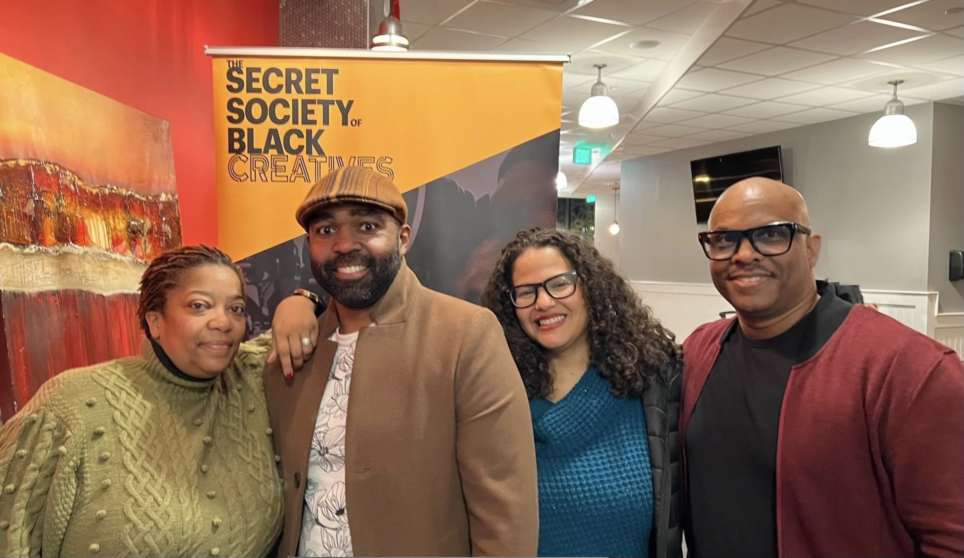 Four people, Award Recipients of the Common Good Award, smile together in front of a yellow sign that reads The Secret Society of Black Creatives in a modern, well-lit room with art on the walls.