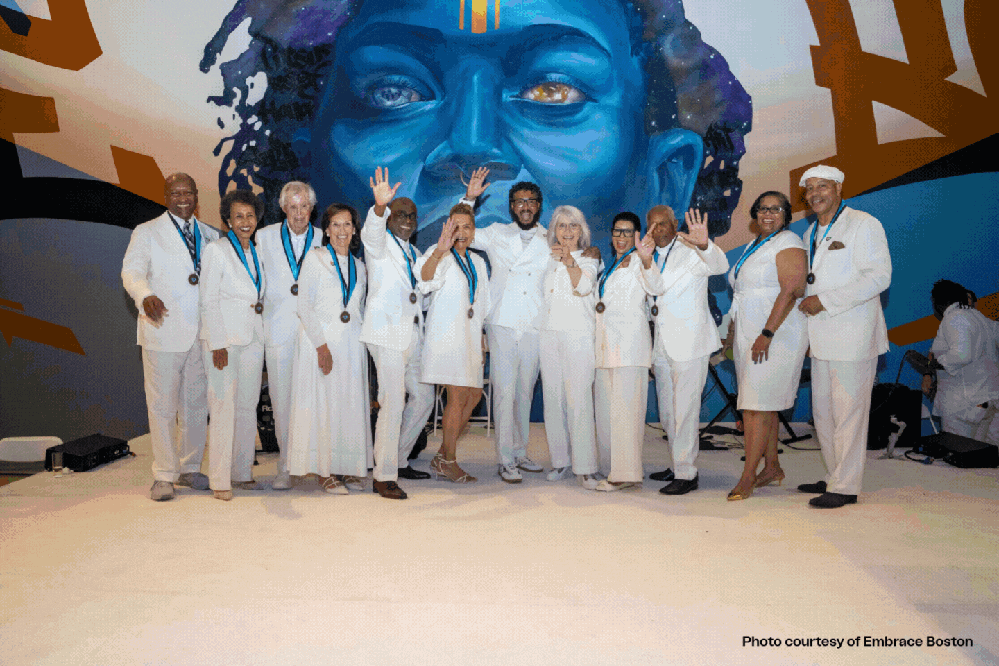 A group of people dressed in white pose together on stage, smiling and waving, all wearing medals. Behind them is a large mural of a blue-toned face. Photo courtesy of Embrace Boston for the MassArt Newsletter Summer 2025 edition.