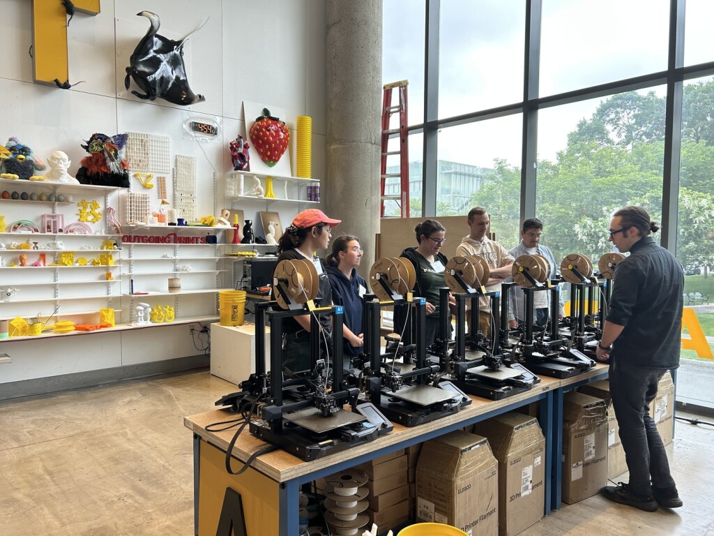 A group of people stands by a row of 3D printers in a bright, modern workshop filled with colorful art and objects on shelves, while an instructor speaks to them. Large windows let in natural light.