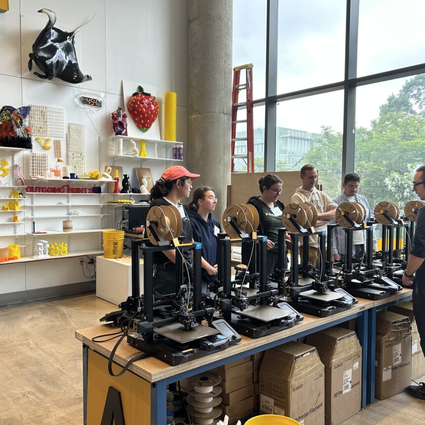 A group of people stands by a row of 3D printers in a bright, modern workshop filled with colorful art and objects on shelves, while an instructor speaks to them. Large windows let in natural light.