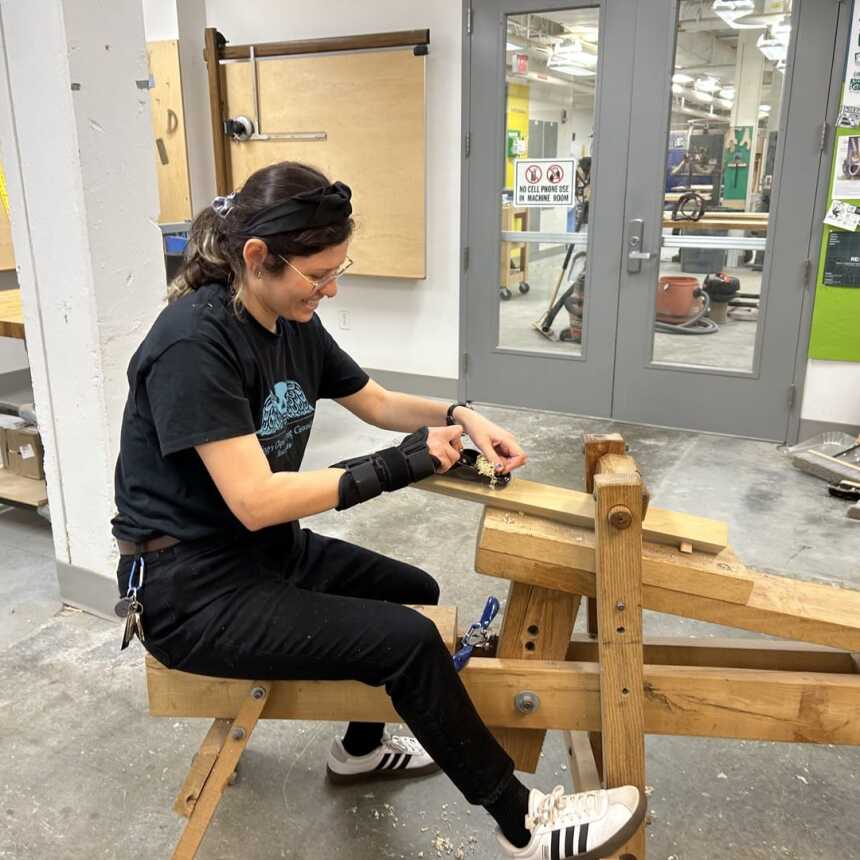 A person wearing safety glasses and a black outfit uses a woodworking tool on a piece of wood, seated on a wooden workbench in a workshop with tools and posters on the walls.