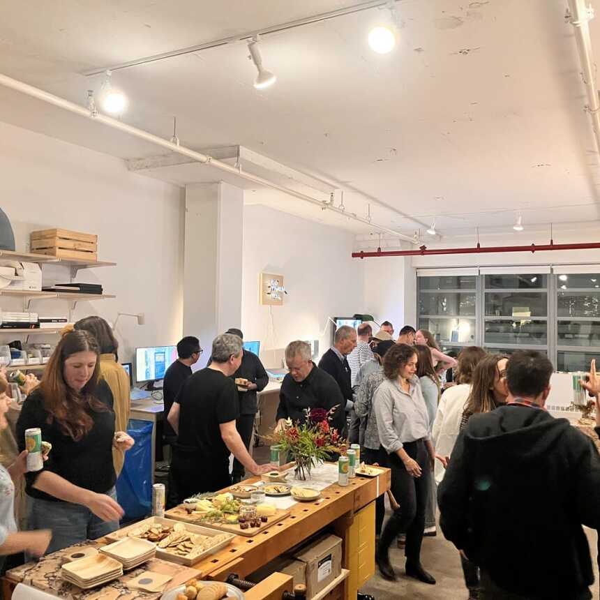 A group of people socialize and eat around a table with snacks and drinks in a bright, modern indoor space with shelves, computers, and large windows. The atmosphere is lively and casual.