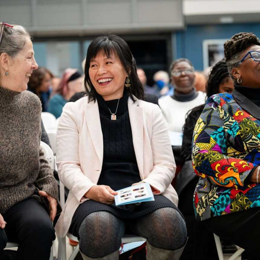 Three women sit together in a lively crowd, smiling and laughing. One wears a tan sweater, another a white blazer, and the third a colorful patterned jacket. They appear to be enjoying an event or presentation.