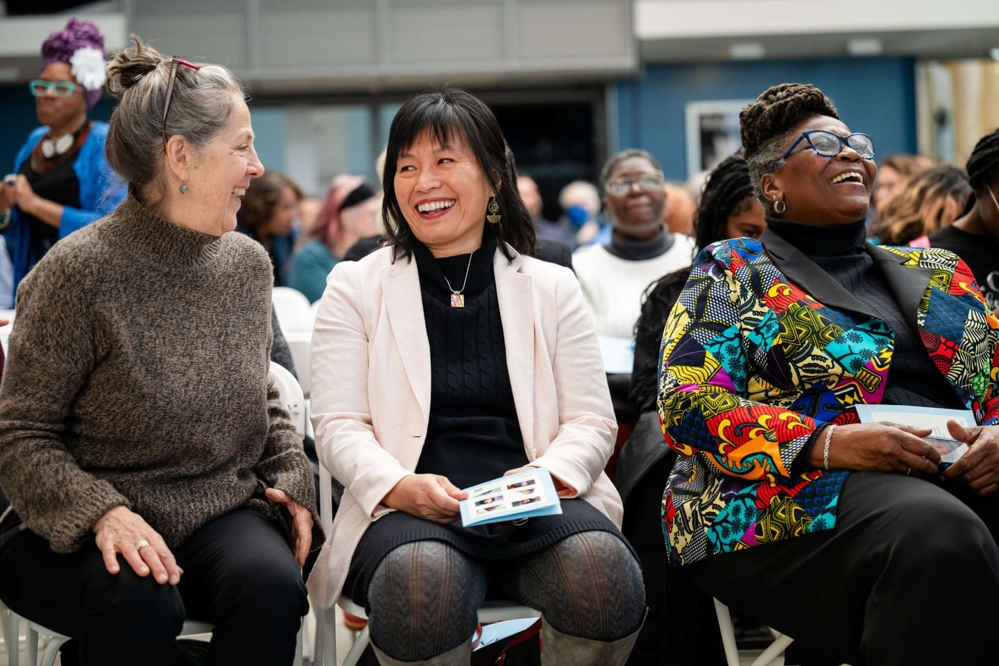 Three women sit together in a lively crowd, smiling and laughing. One wears a tan sweater, another a white blazer, and the third a colorful patterned jacket. They appear to be enjoying an event or presentation.