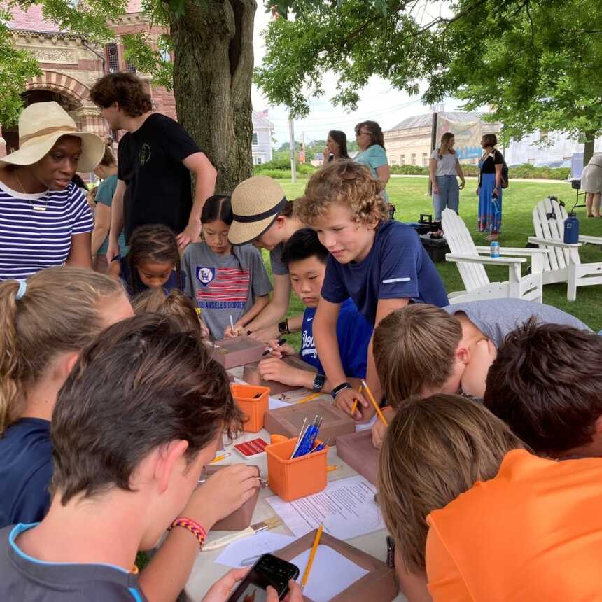 A group of children gather around a table outdoors, working on activities with pencils and paper. Adults and other kids stand nearby under a tree, and white chairs are set up on the grass in the background.