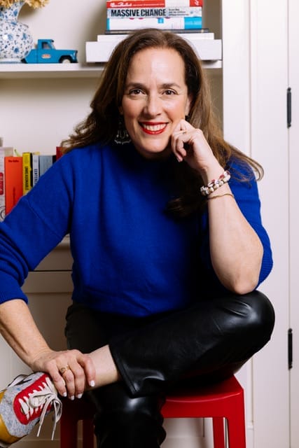 A woman with long brown hair, wearing a bright blue sweater, black leather pants, and gray sneakers with red laces, sits on a red stool, smiling and resting her chin on her hand in a cozy room filled with books, decor, and MassArt Board of Trustees materials.