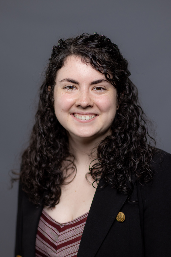 A person with long, curly dark hair is smiling at the camera. They are wearing a black blazer over a maroon and white striped top, standing against a plain gray background.
