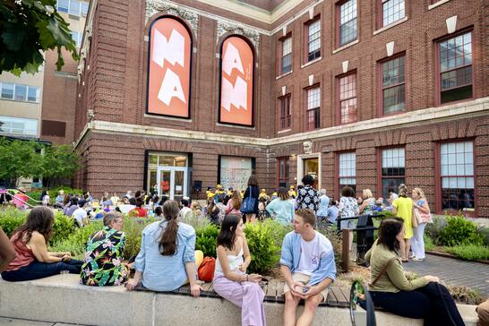 A diverse group of people gather outside the large brick MassArt museum building with tall windows and banners reading MAM. Some chat, while others walk or relax on benches, enjoying a lively outdoor event.