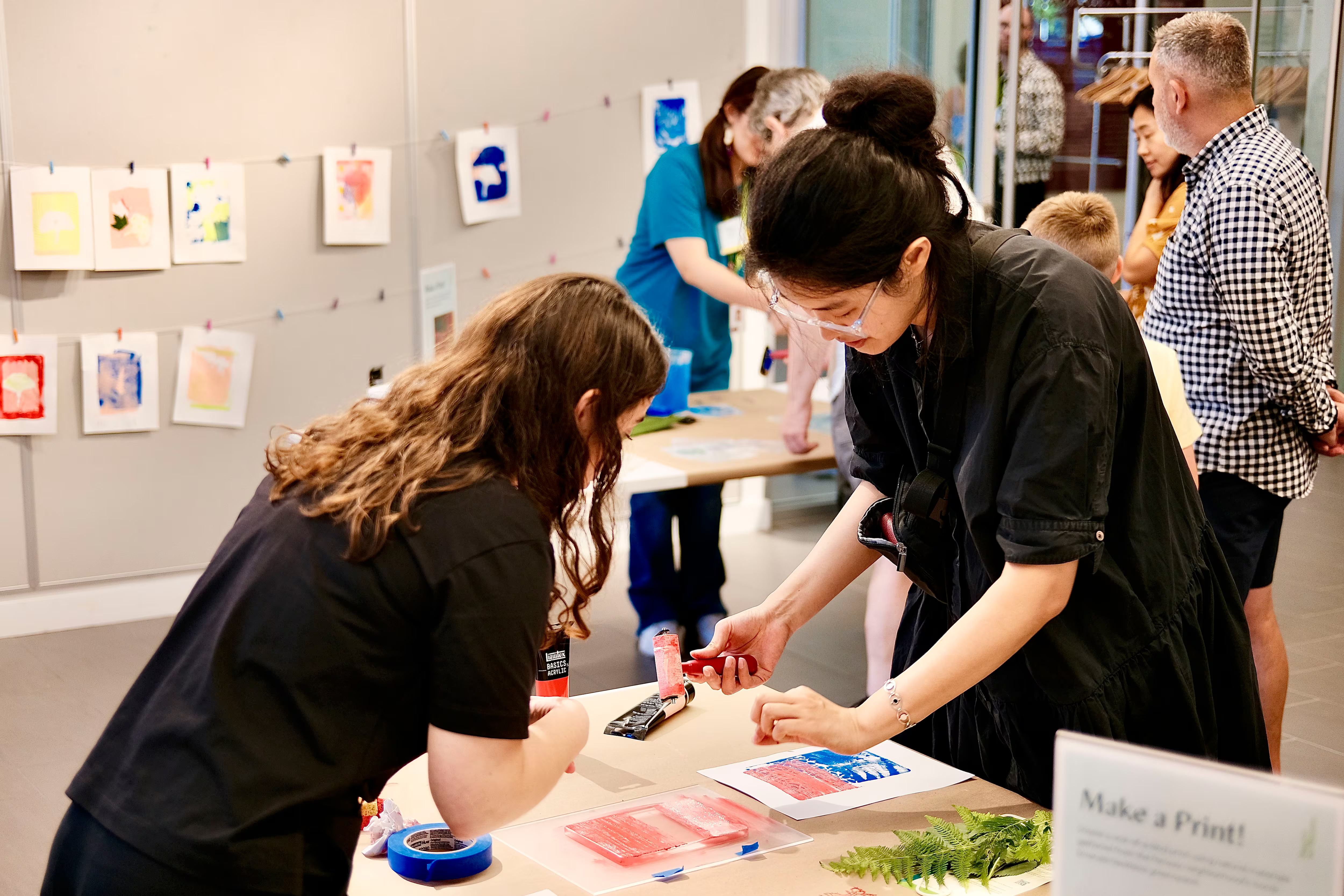 People participate in an art workshop at MassArt Art Museum, working with printmaking materials at tables. Art prints hang on the wall, and supplies are spread out as adults and a child enjoy Best of the Best creative activities in a bright room.