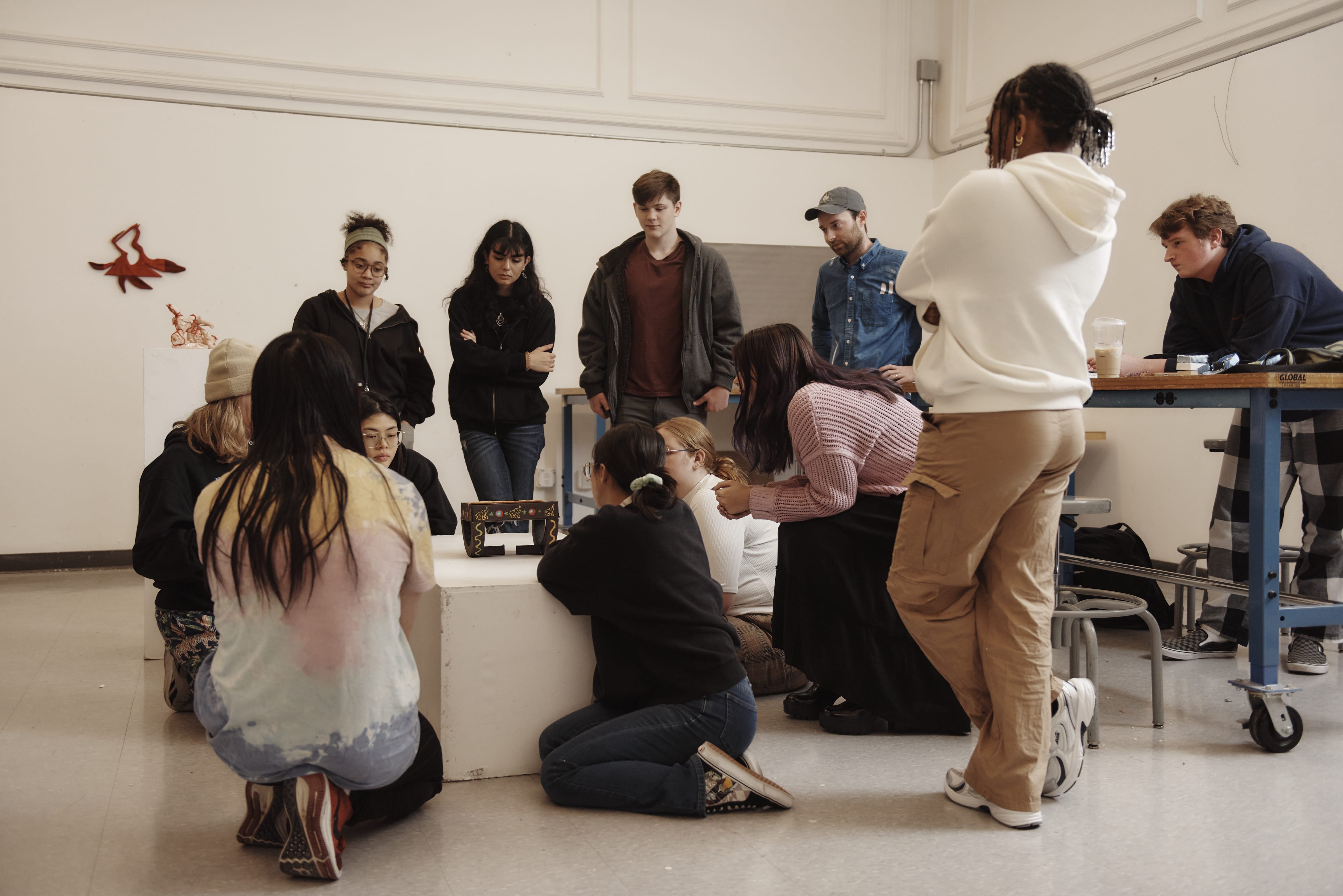 A diverse group of students at MassArt gathers in a classroom, some standing and others kneeling, focused on a small display on a white pedestal, with art supplies and wall decorations visible—reflecting why its listed among Americas Best Colleges by Money Magazine.