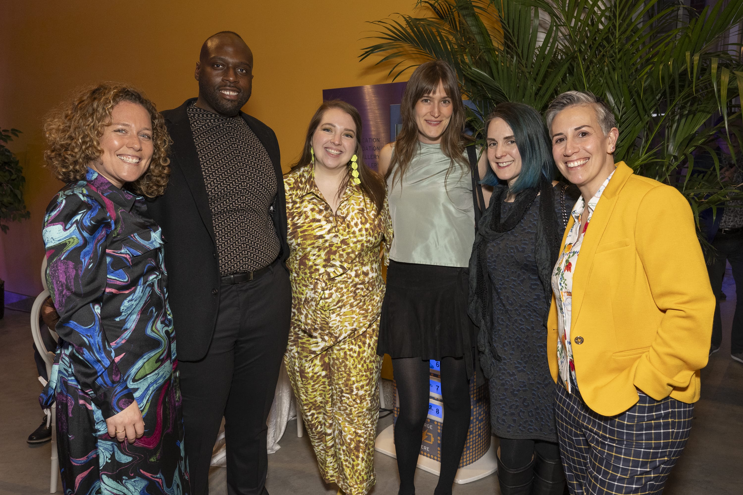 Six people from the Auction Committee stand together indoors, smiling at the camera in colorful, stylish outfits. Tall green plants and a yellow wall in the background create a warm, lively atmosphere.
