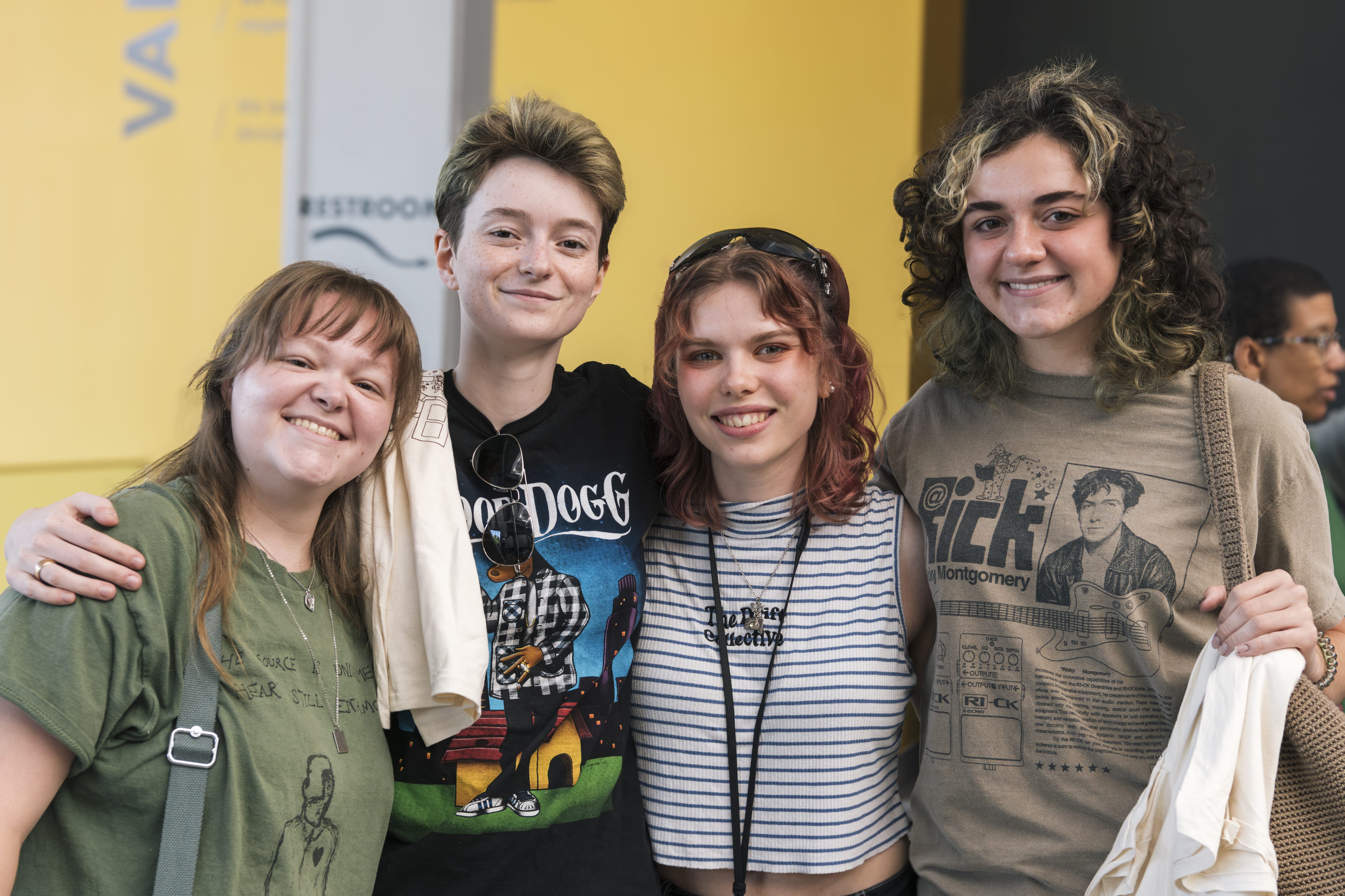 Four smiling young adults stand close together, posing for a photo. They wear casual clothing, including graphic tees, against a yellow and black background. They appear happy and are enjoying each others company.
