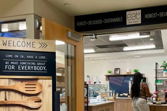 A woman stands at the counter inside a small modern food market with a sign reading eat Food Market: Honest - Delicious - Sustainable. A welcome sign with wooden utensils invites dining near the entrance.