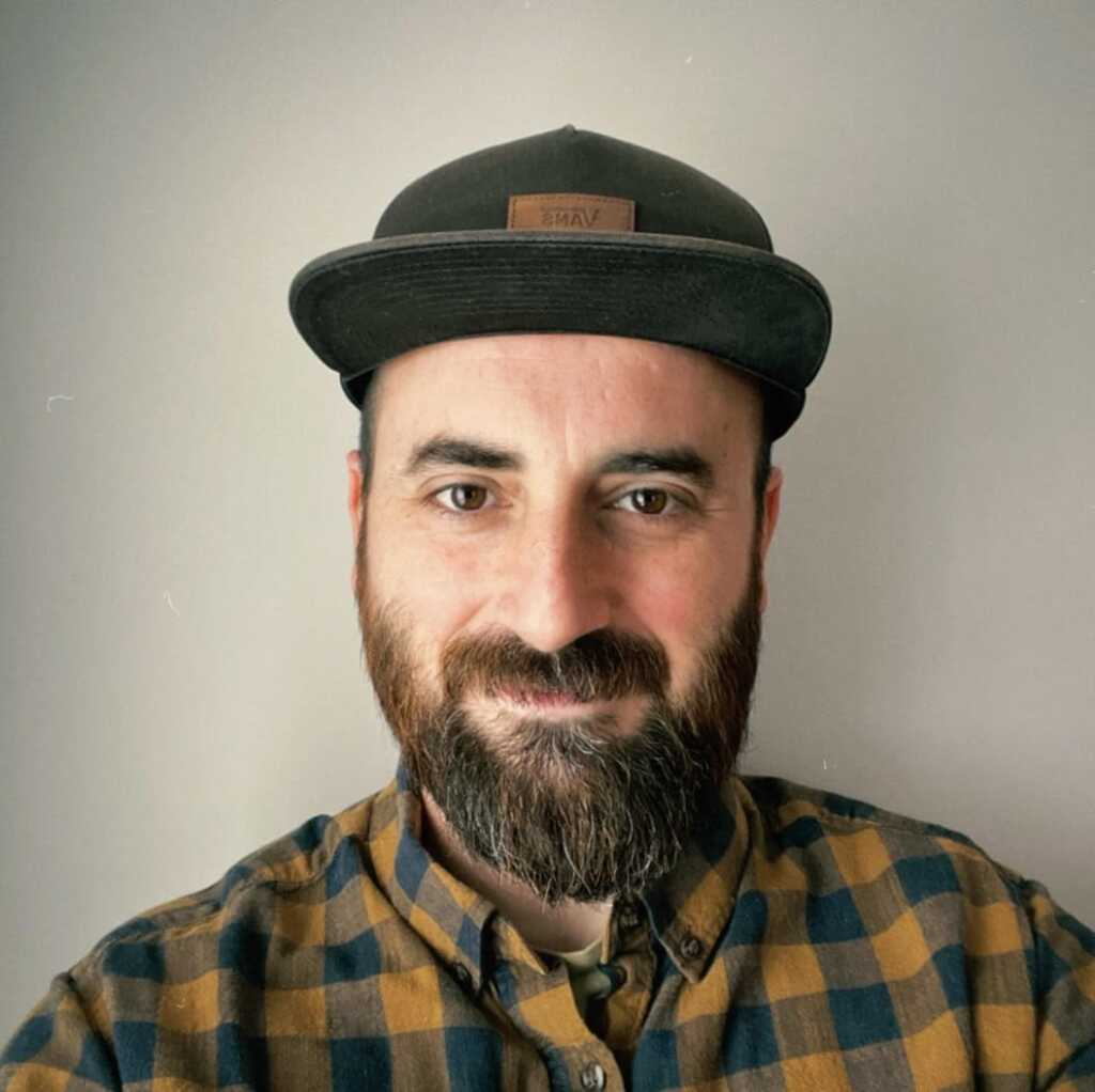 A man with a beard and mustache smiles at the camera. He is wearing a black cap and a yellow and brown plaid shirt, standing indoors against a plain, light-colored background.