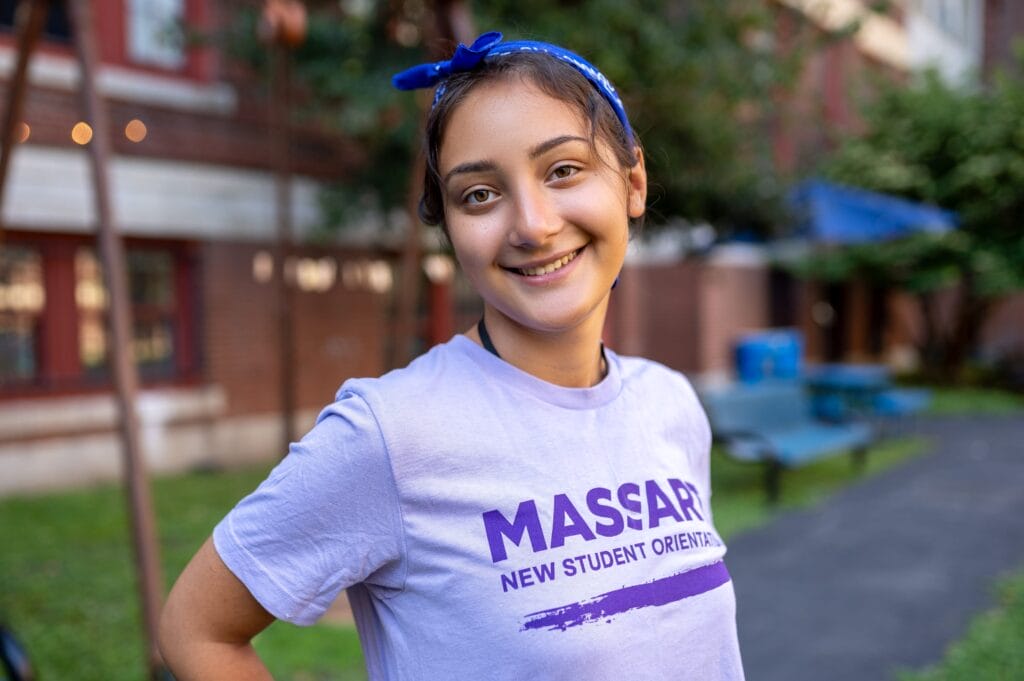 A smiling young woman, Kailey Caballero, stands outdoors in a courtyard, wearing a purple MassArt New Student Orientation t-shirt and a blue headband—embracing campus life and finding your voice amid brick buildings and greenery.