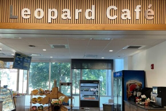 Leopard Café interior with a wooden sign above, snacks and drinks displayed on shelves and racks, a refrigerated section, pastry case, and large windows providing natural light for a cozy dining experience in the background.