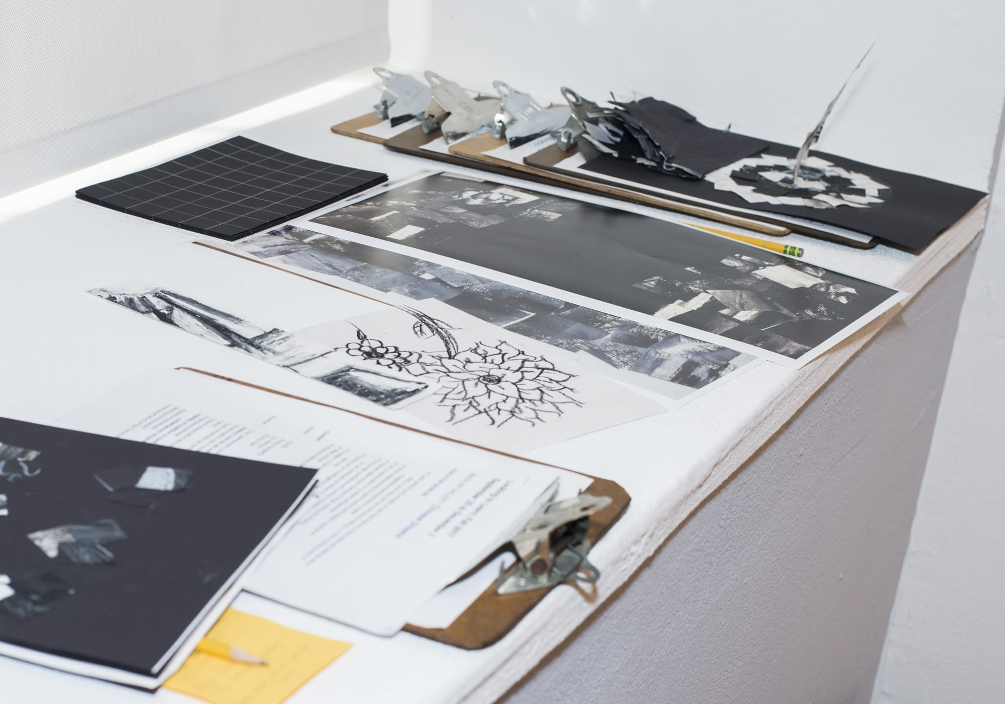 A tabletop displaying an art portfolio with clipboards, papers featuring black-and-white photos, a grid notebook, metal bulldog clips, and abstract sketches—all neatly arranged in a bright, well-lit space.