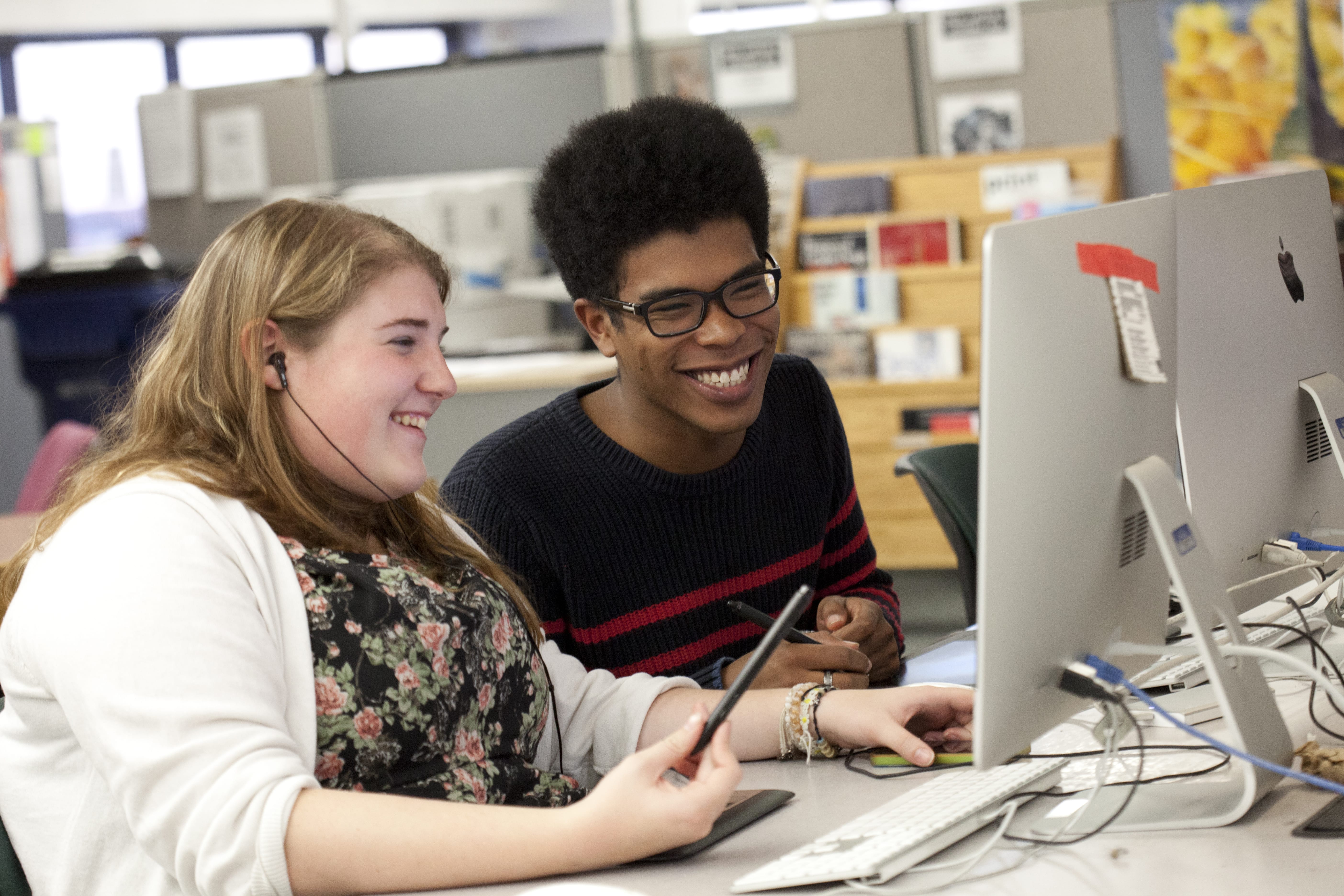 Two students sitting side by side at a desk, smiling and working together on desktop computers. One holds a stylus, and both appear engaged and happy in a classroom or computer lab setting.