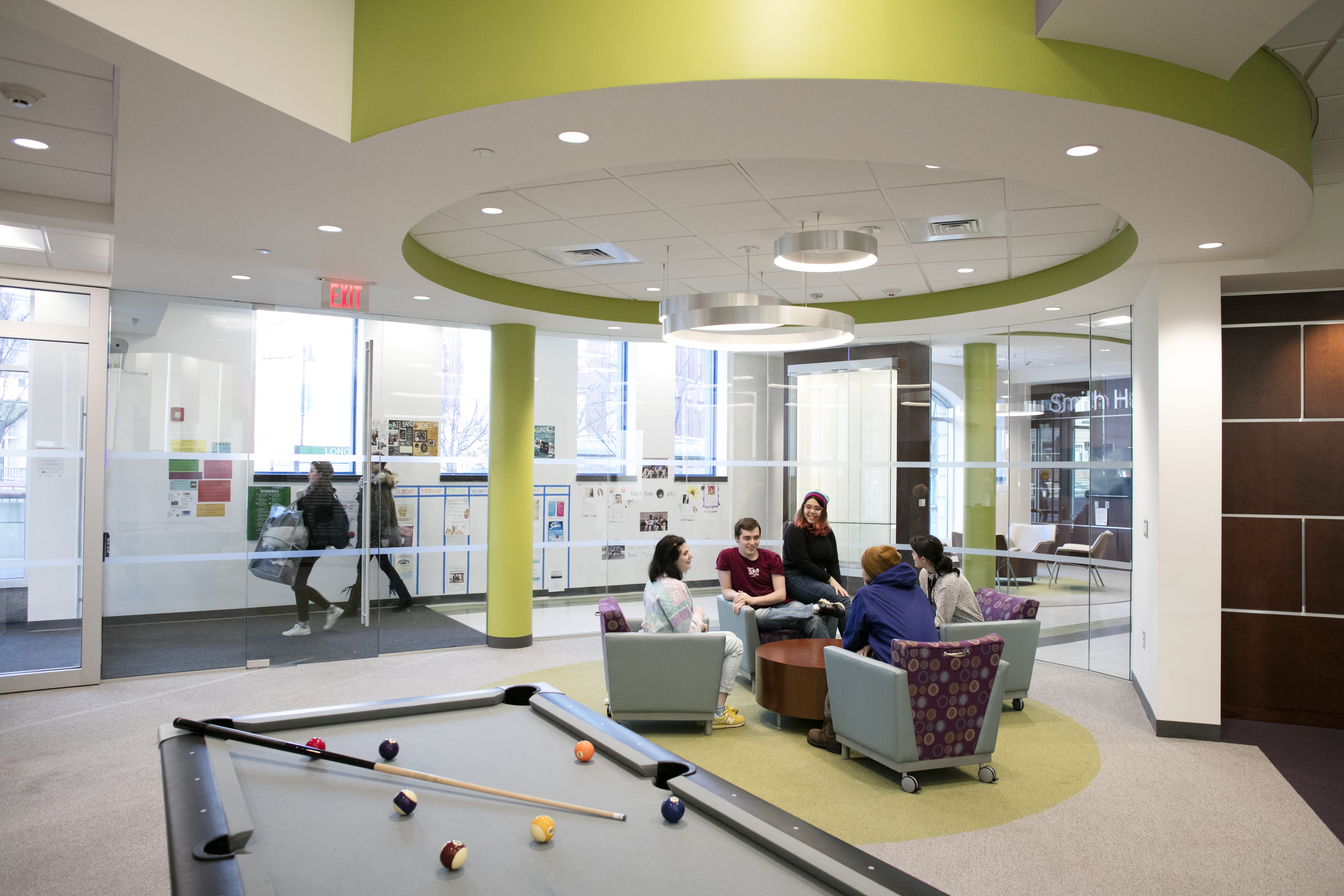 A group of six new students sit and talk in a modern lounge area with purple chairs and a round table. A pool table is in the foreground, and large windows line the background, letting in natural light.