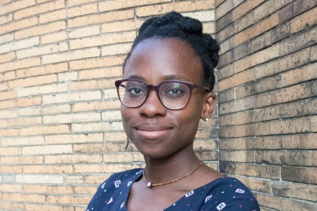 A member of the Alumni Leadership Council, a woman with glasses and braided hair stands smiling with crossed arms, wearing a patterned navy blue dress, in front of a brick wall.