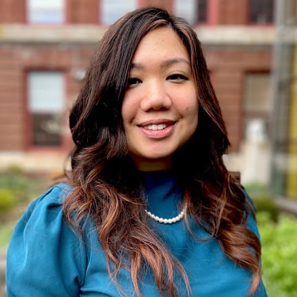 A woman with long wavy hair wearing a blue top and a pearl necklace smiles outdoors in front of a brick building.