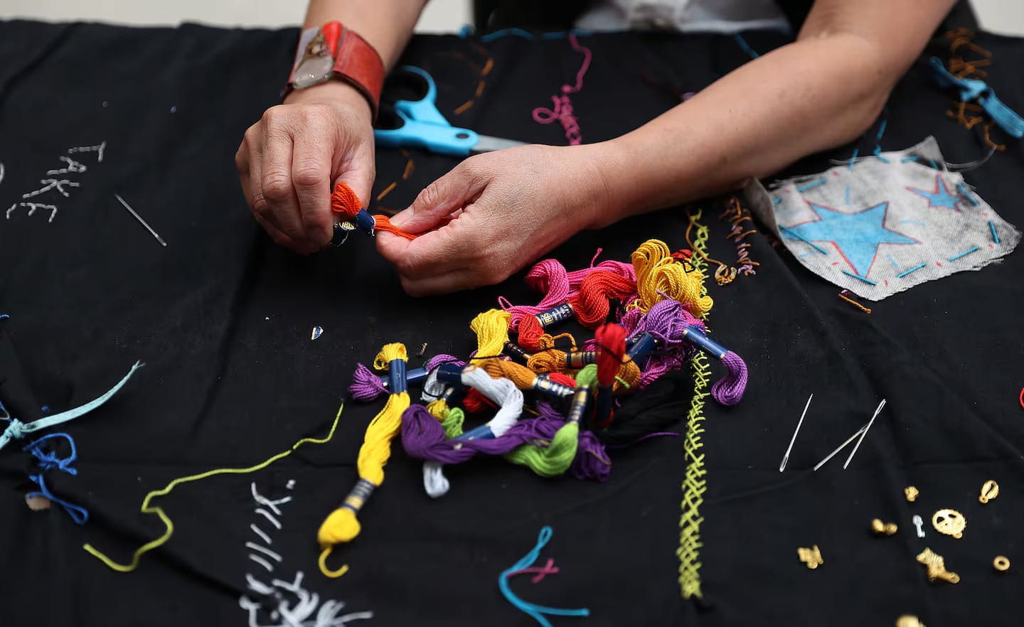 A person arranges colorful embroidery threads on a black fabric, surrounded by needles, a pair of blue scissors, and stitched designs, including stars and zigzag patterns.