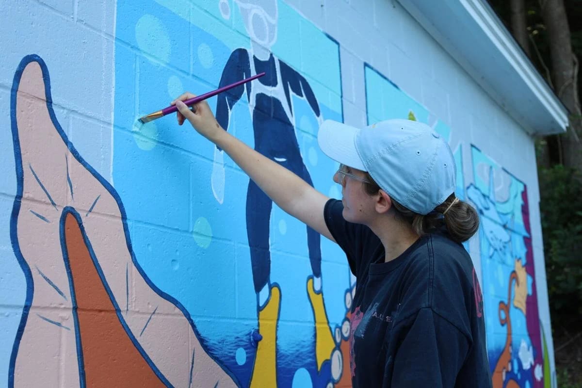 A MassArt Alum wearing a light blue cap and glasses paints a colorful underwater mural featuring divers and marine life on an outdoor wall at Assabet Park in Northborough.