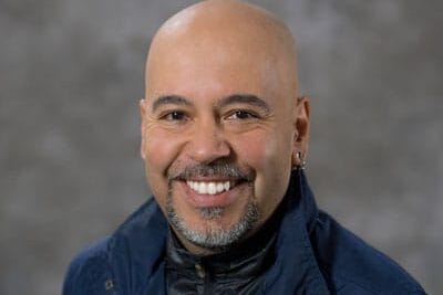 A smiling bald man with a goatee and earrings, wearing a navy blue jacket over a black zip-up top, poses in front of a gray, blurred background—proudly representing the Alumni Leadership Council.