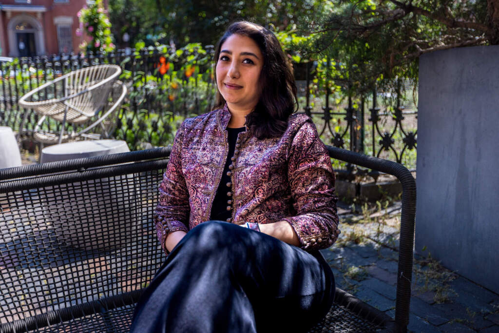 A woman with dark hair sits on an outdoor bench, wearing a patterned jacket and dark pants, with one leg crossed over the other. Greenery and a wrought iron fence are visible in the background.