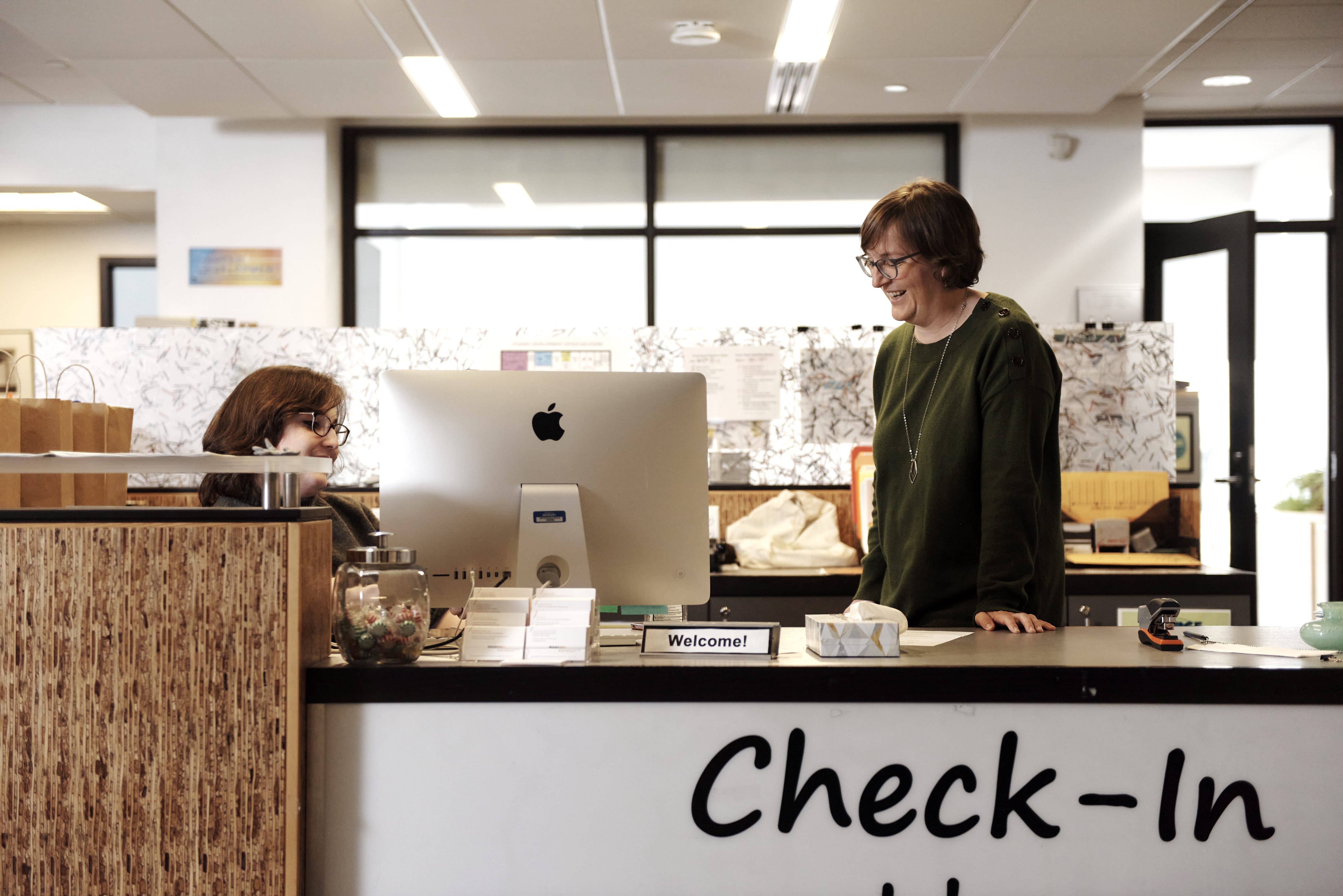 Two people interact at a check-in desk in a modern office. One provides academic support from behind an iMac computer, while the other stands, smiling, across the counter. The desk displays a Welcome! sign and a glass jar.