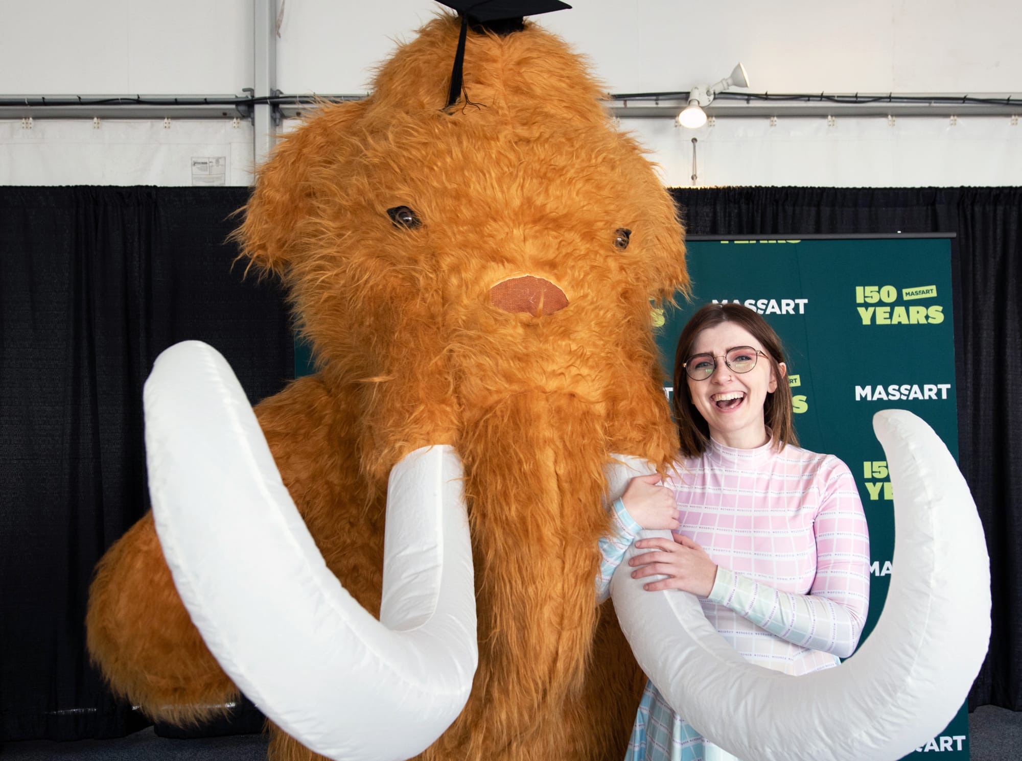 A smiling person stands next to a mammoth mascot with large tusks and a graduation cap at a MassArt 150th anniversary event, hosted by the Office of Student Engagement. A step-and-repeat banner appears in the background.