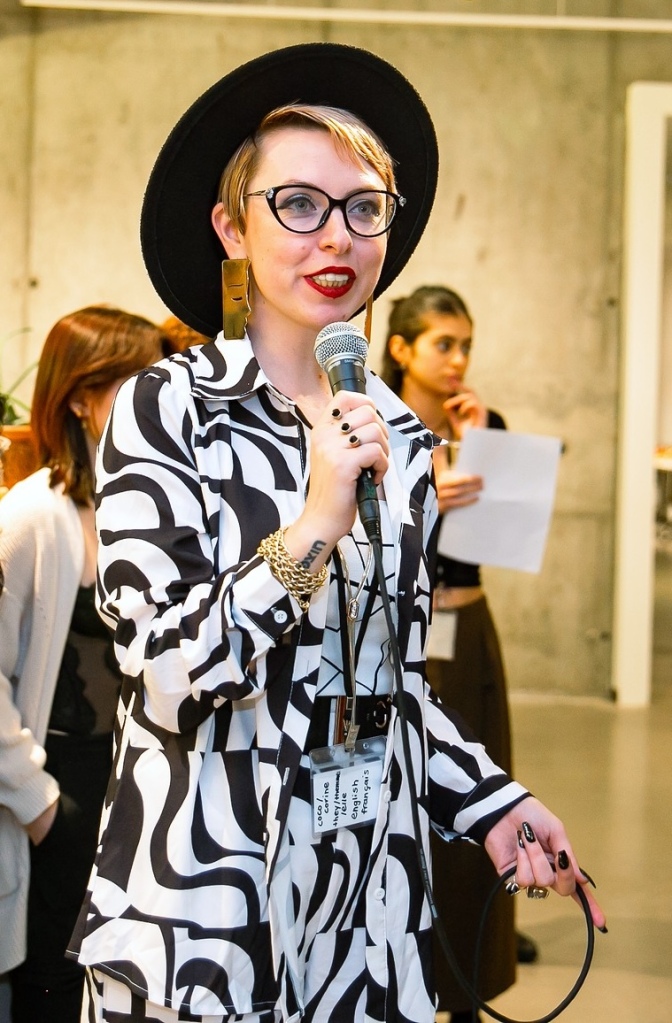 A person wearing glasses, a black hat, and a black-and-white patterned outfit holds a microphone while speaking. Several people stand in the background in an indoor setting.
