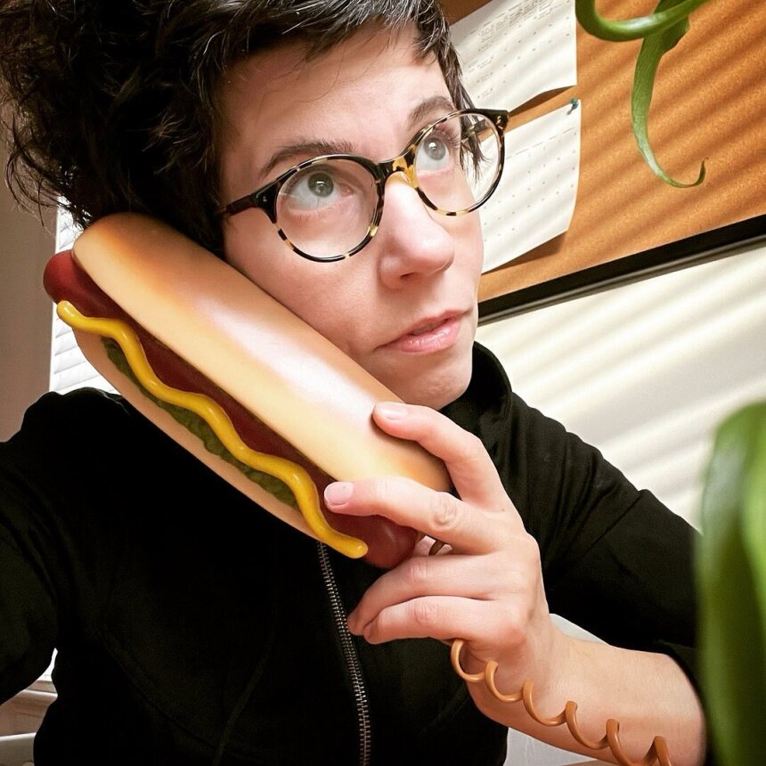 A person with short dark hair and glasses holds a telephone shaped like a hot dog to their ear, looking thoughtful. The scene is indoors with natural light and a plant visible in the foreground.