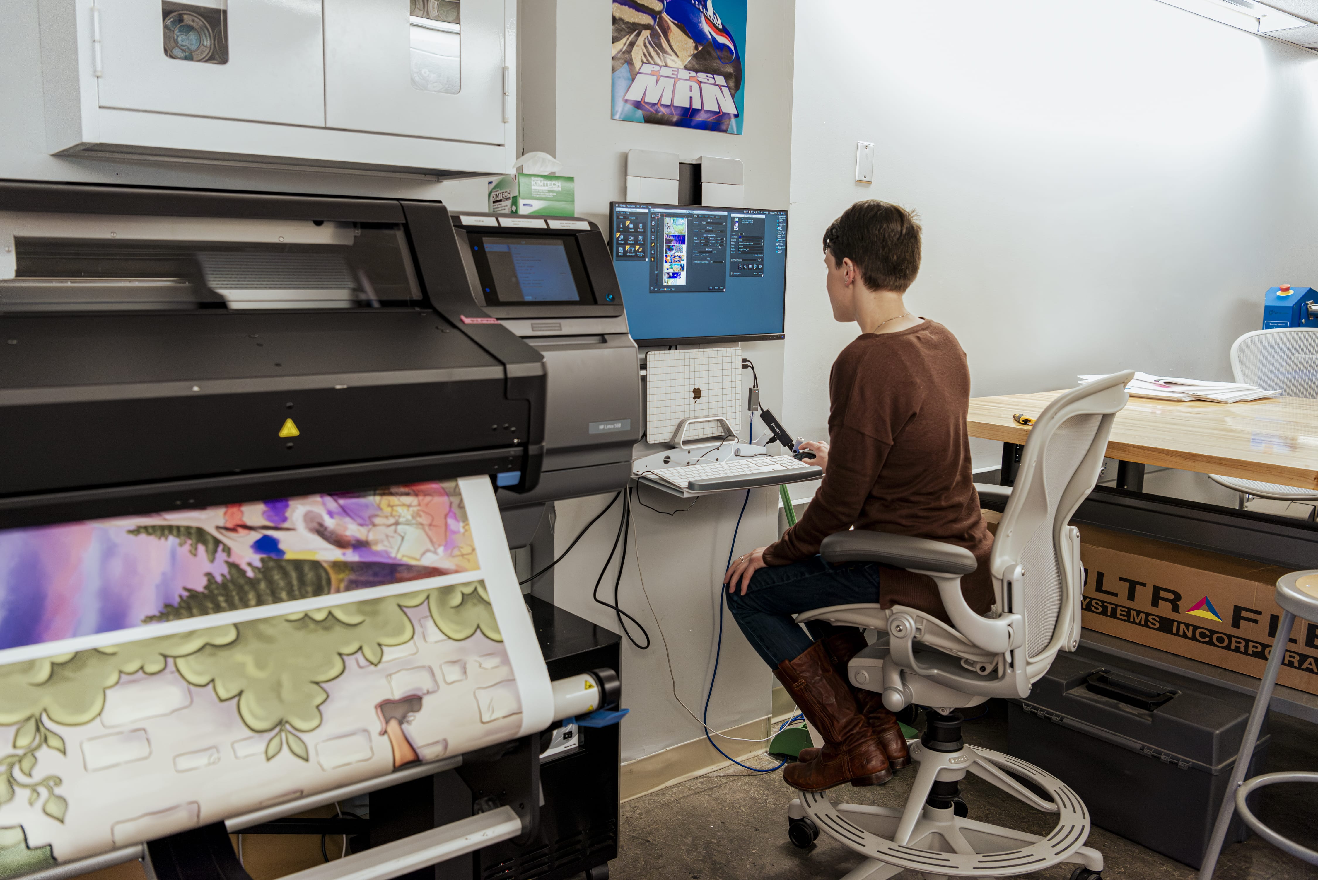 A person sits at a desk using a computer in a print shop. A large printer beside them is printing a colorful image, and the workspace is filled with equipment and supplies.