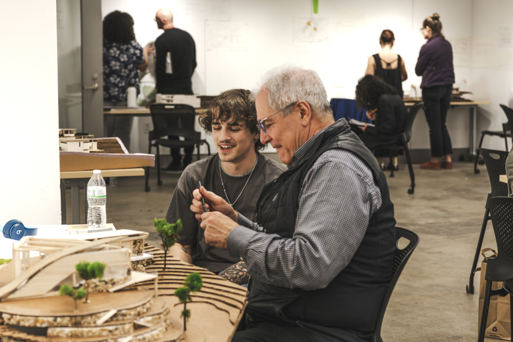 An older man and a young man sit at a table, smiling and working on a model project together in a classroom, highlighting the value of academic support. Other people are collaborating and working in the background.