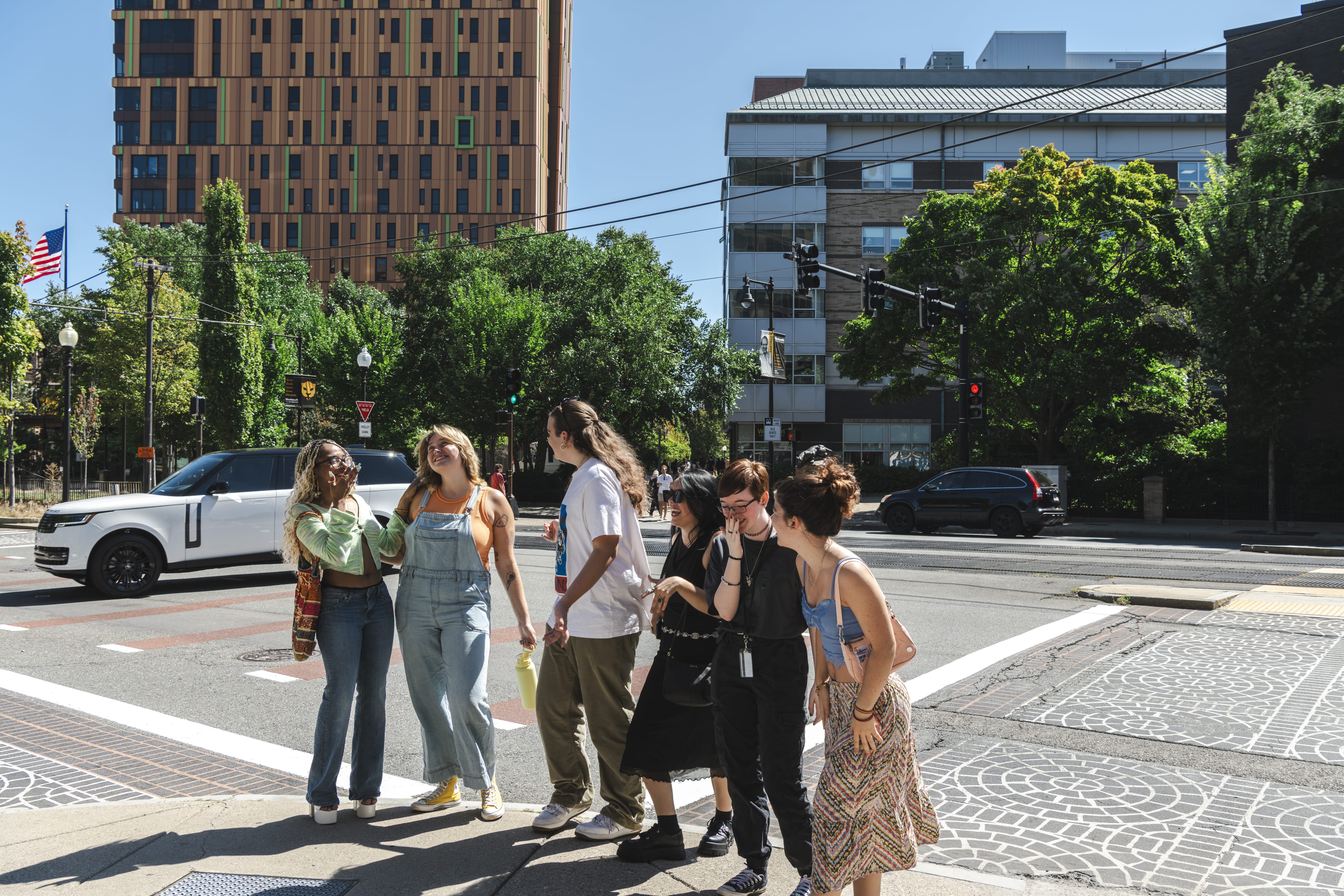 A diverse group of seven people stands together at a crosswalk on a sunny day, chatting and laughing, with modern buildings and greenery in the background.