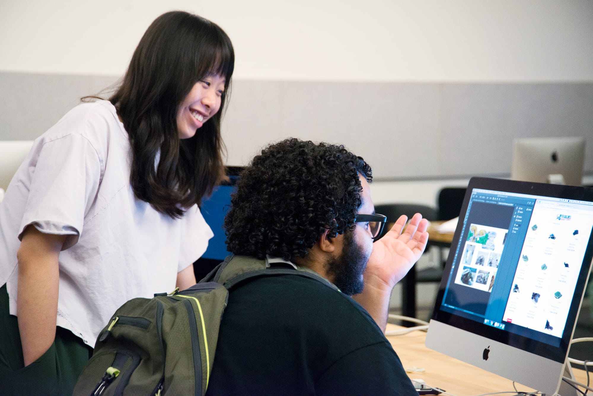 A woman smiles while standing next to a man seated at a computer. The man gestures towards the screen, which displays various images, as they enjoy a friendly conversation about academic support in a classroom or office setting.
